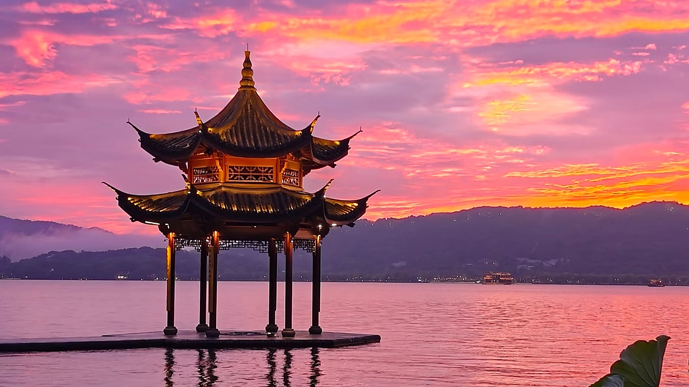 Leifeng Pagoda reflected in West Lake in Hangzhou, Zhejiang province, with the multi-tiered golden-brick pagoda rising above green summer foliage and lotus leaves on the calm water surface of the lake in soft afternoon light