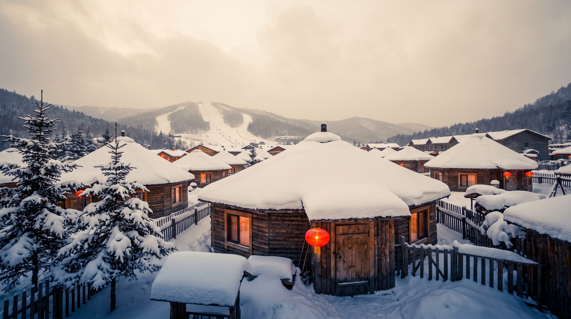 China Snow Town at night with red lanterns glowing against snow-covered wooden houses, snow piled high on rooftops under a deep blue winter sky